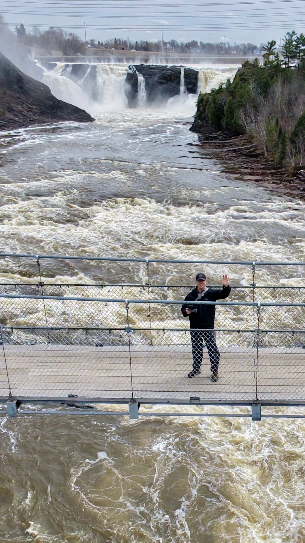Vue aérienne de la passerelle suspendue du parc des Chutes-de-la-Chaudière à Lévis, une personne salue le drone, la chute en arrière-plan